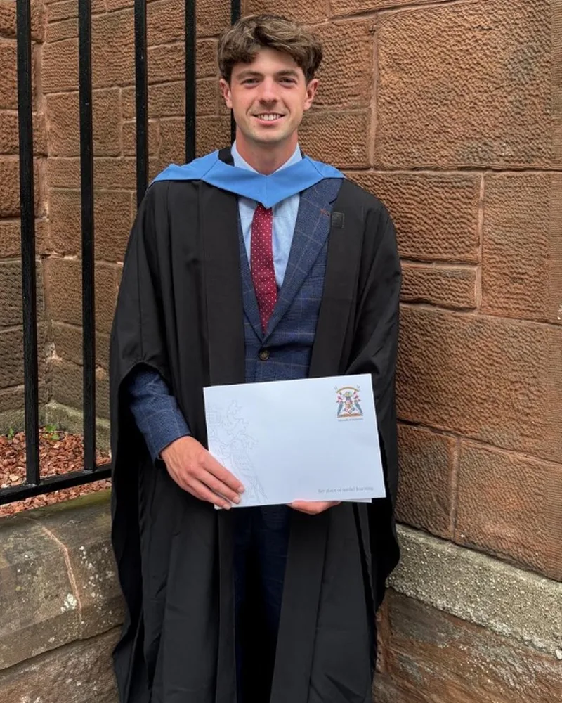 Douglas Stark in graduation gown and hood, holding his degree outside a red sandstone building
